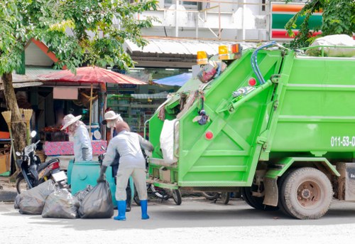 A green rubbish collection truck is parked on a street, positioned adjacent to a curb in an urban area. The truck's rear compaction section is open, exposing various waste compartments, while a worker dressed in grey overalls, a white hat, and blue gloves is actively loading black garbage bags into the rear section of the vehicle. Several black refuse bags are placed on the pavement nearby, some partially in motion as the worker handles them. In the background, there are small shops with awnings, including a street vendor's stall covered by a pink canopy and other storefronts, with a few pedestrians and parked motorcycles also visible. The scene suggests a routine rubbish collection in a town, with leafy trees providing some shade and ambient daylight illuminating the scene, highlighting the textures of the waste bags, the vehicle's metallic surface, and the surrounding urban environment. The image conveys a typical rubbish removal operation relevant to local waste management services in Upminster or nearby towns.
