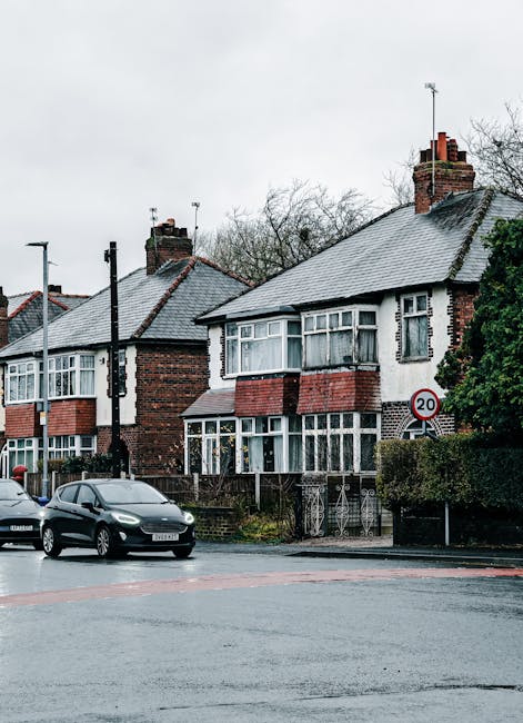 A row of traditional semi-detached houses with brick and white exterior walls, large bay windows, and tiled roofs, situated along a residential street. In the foreground, there are parked cars, includ