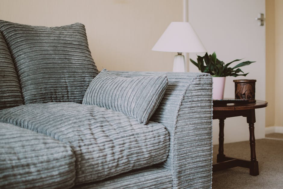 A man with dark curly hair and a beard, dressed in a dark blue uniform, is lifting a small green velvet sofa with a tufted backrest and wooden legs, seeming to prepare for removal. The sofa is positio