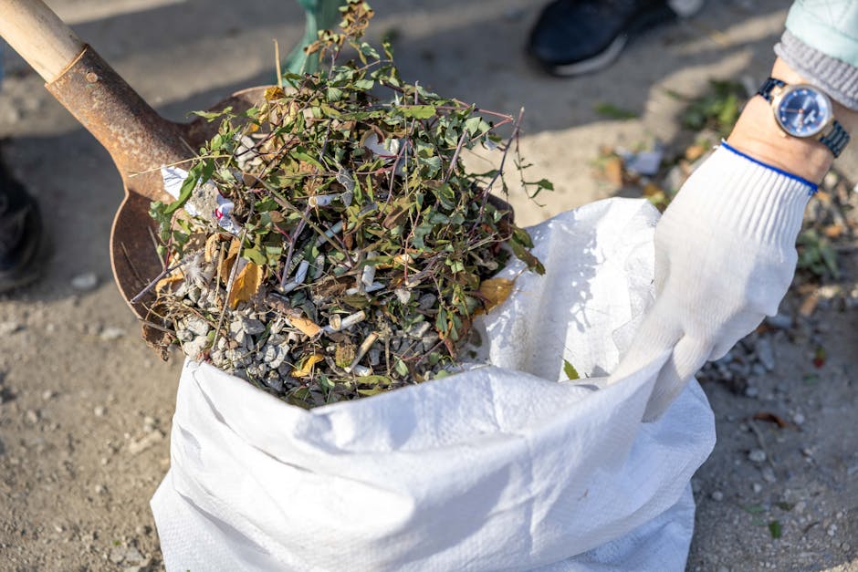A close-up photograph of a person wearing a white glove and a watch, holding a white woven polypropylene sack filled with garden debris. The debris consists of small leafy branches, twigs, and gravel-
