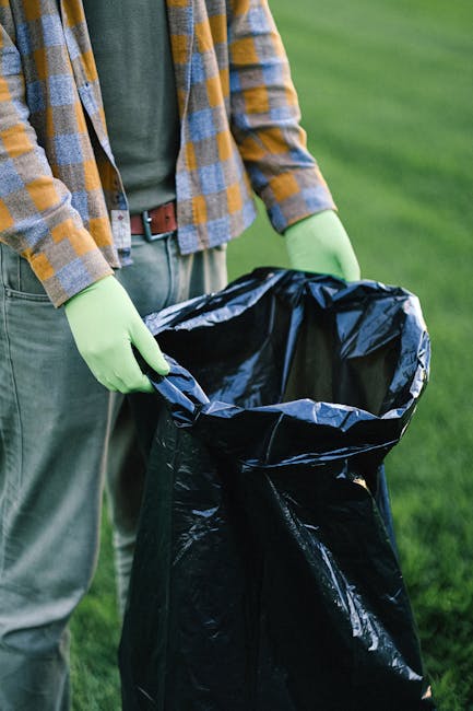 A close-up photograph of a person wearing a white glove and a watch, holding a white woven polypropylene sack filled with garden debris. The debris consists of small leafy branches, twigs, and gravel-